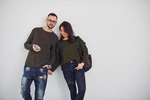 Cheerful multi ethnic couple with backpack and phone standing together indoors in the studio against white background photo