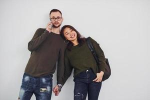 Cheerful multi ethnic couple with backpack and phone standing together indoors in the studio against white background photo