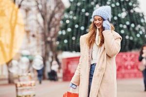 Young girl in warm clothes with shopping bags in hand have a walk outdoors in the city and holding phone photo