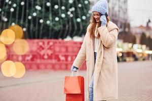 Young girl in warm clothes with shopping bags in hand have a walk outdoors in the city and holding phone photo