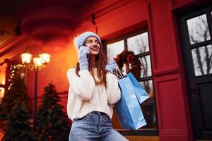 una joven con ropa de abrigo da un paseo al aire libre en la ciudad por la noche cerca del edificio con luces festivas artificiales. hablando por telefono foto