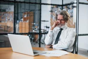 aburrido hombre de negocios maduro con cabello gris y barba en ropa formal está en la oficina contra la computadora portátil foto