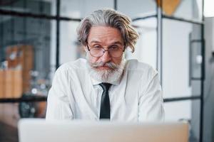 Expressive mature businessman with grey hair and beard in formal clothes is in the office against laptop photo