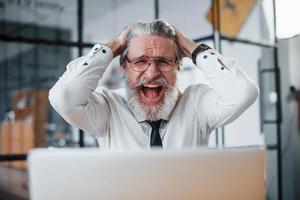 Expressive mature businessman with grey hair and beard in formal clothes is in the office against laptop photo