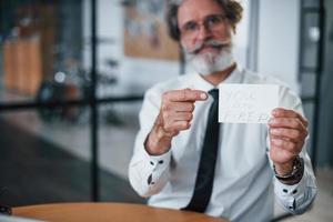 Holding paper with text you are fired. Mature businessman with grey hair and beard in formal clothes is in the office photo