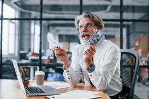 Happy mature businessman with grey hair and beard in formal clothes is in the office holding money photo