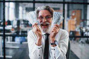 Happy mature businessman with grey hair and beard in formal clothes is in the office holding money photo