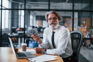 Happy mature businessman with grey hair and beard in formal clothes is in the office holding money photo