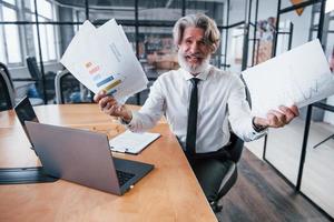 Holding documents in hands. Mature businessman with grey hair and beard in formal clothes is in the office photo