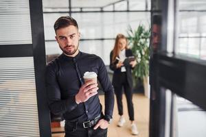 Modern businessman standing indoors in office. His female colleague behind photo