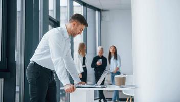 Man in formal clothes standing and reading documents in front of young successful team that working and communicating together indoors in office photo