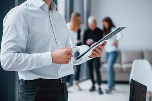 Man in formal clothes standing with documents in front of young successful team that working and communicating together indoors in office photo