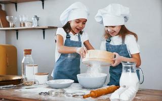 dos niñas pequeñas con uniforme de chef azul trabajando con harina usando tamiz en la cocina foto