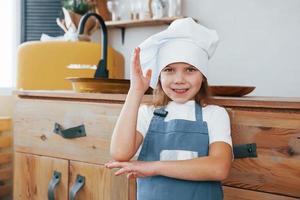 linda niñita con sombrero blanco y uniforme azul parada en el interior de la cocina foto