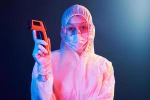 Nurse in mask and white uniform standing in neon lighted room and holding infrared thermometer. Stop coronavirus spreading photo