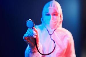 Nurse in mask and white uniform and with stethoscope standing in neon lighted room photo