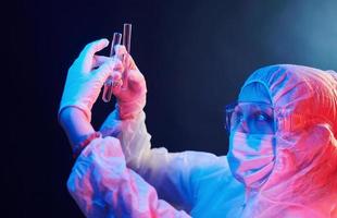 Nurse in mask and white uniform standing in neon lighted room and holding tubes with samples photo