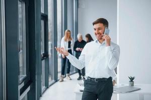 Man in formal clothes talking by phone and standing in front of young successful team that working and communicating together indoors in office photo