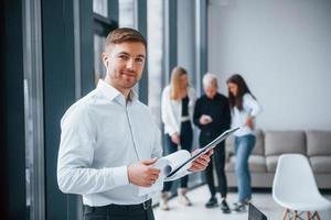 Man in formal clothes standing with documents in front of young successful team that working and communicating together indoors in office photo