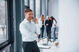 Man in formal clothes talking by phone and standing in front of young successful team that working and communicating together indoors in office photo