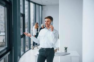 Man in formal clothes talking by phone and standing in front of young successful team that working and communicating together indoors in office photo