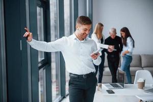 Man in formal clothes with phone and in headphones standing in front of young successful team that working and communicating together indoors in office photo