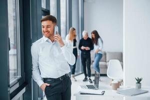 Man in formal clothes talking by phone and standing in front of young successful team that working and communicating together indoors in office photo