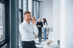 Man in formal clothes talking by phone and standing in front of young successful team that working and communicating together indoors in office photo