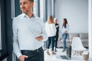 Man in formal clothes standing with phone in hand in front of young successful team that working and communicating together indoors in office photo