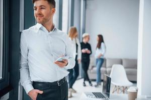 Man in formal clothes standing with phone in hand in front of young successful team that working and communicating together indoors in office photo