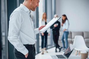 Man in formal clothes standing and reading documents in front of young successful team that working and communicating together indoors in office photo
