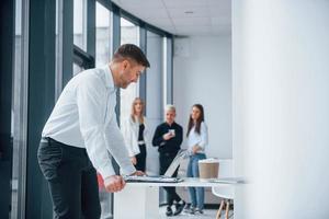 Man in formal clothes standing and reading documents in front of young successful team that working and communicating together indoors in office photo