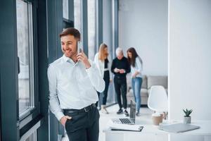 Man in formal clothes talking by phone and standing in front of young successful team that working and communicating together indoors in office photo