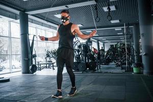 un joven fuerte con ropa deportiva y una máscara aterradora haciendo ejercicio usando una cuerda para saltar en el gimnasio foto