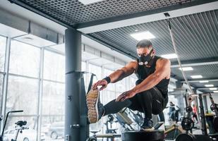 Strong young man in sportive clothes and scary mask doing push ups on one leg in gym photo