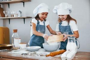 Two little girls in blue chef uniform working with flour by using sieve on the kitchen photo