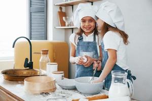 Two little girls in blue chef uniform talking secrets to each other when preparing food on the kitchen photo