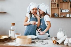 Two little girls in blue chef uniform talking secrets to each other when preparing food on the kitchen photo
