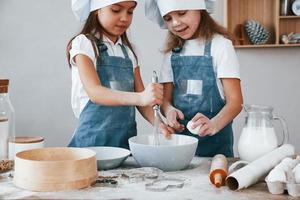dos niñas pequeñas con uniforme de chef azul mezclando harina con huevos en un plato en la cocina foto