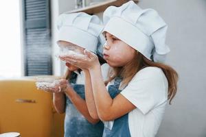 Two little girls in blue chef uniform blows the flour out of hands on the kitchen photo