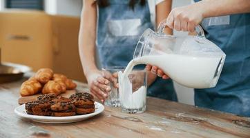 Close up view of two little girls in blue chef uniform that pouring milk into glasses on the kitchen with cookies on table photo
