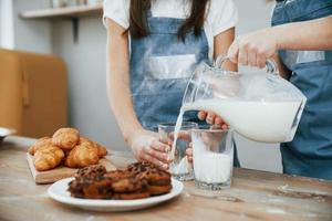 Close up view of two little girls in blue chef uniform that pouring milk into glasses on the kitchen with cookies on table photo