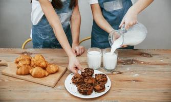 Close up view of two little girls in blue chef uniform that pouring milk into glasses on the kitchen with cookies on table photo