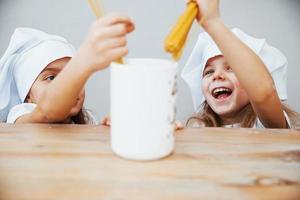 Two cute little girls in white chef hats holding spaghetti and puts it to big cup photo