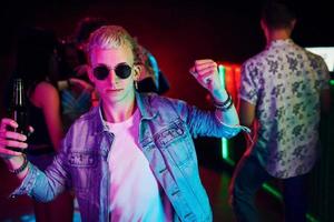 Hipster guy in sunglasses and with bottle of alcohol posing for camera in front of young people that having fun in night club with colorful laser lights photo