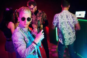 Hipster guy in sunglasses and with bottle of alcohol posing for camera in front of young people that having fun in night club with colorful laser lights photo