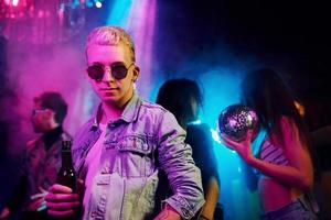 Hipster guy in sunglasses and with bottle of alcohol posing for camera in front of young people that having fun in night club with colorful laser lights photo