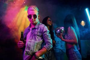 Hipster guy in sunglasses and with bottle of alcohol posing for camera in front of young people that having fun in night club with colorful laser lights photo