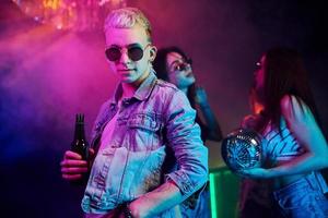 Hipster guy in sunglasses and with bottle of alcohol posing for camera in front of young people that having fun in night club with colorful laser lights photo