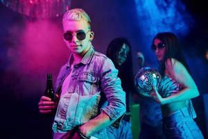 Hipster guy in sunglasses and with bottle of alcohol posing for camera in front of young people that having fun in night club with colorful laser lights photo